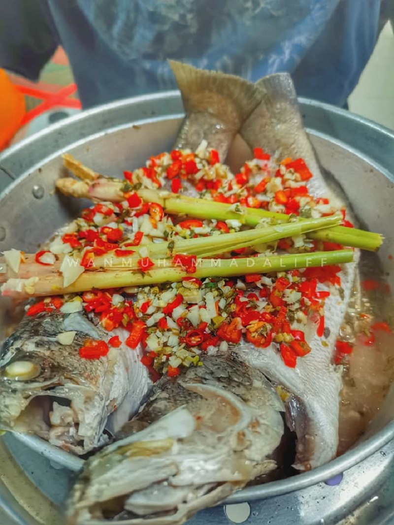 Ikan siakap kukus bersama limau nipis dan daun ketumbar dalam pinggan.